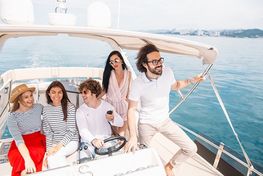 Young curly man navigating a seaboat, sailing in company of his joyful friends, guys and girls sitting beside, enjoying fresh sea wind