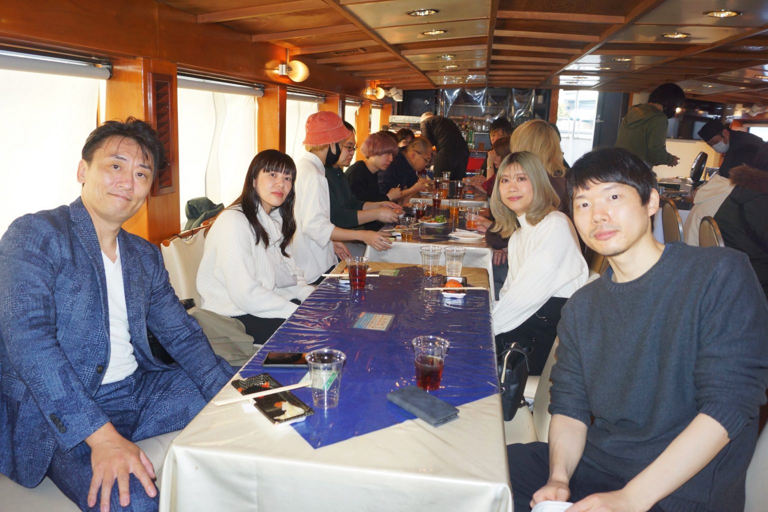 船内で着席し、グループ写真を撮影する男女。後ろには寿司職人が寿司を握っている。